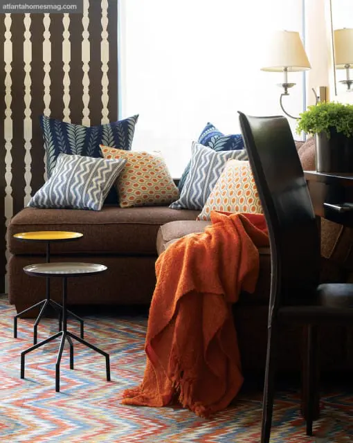 Brown sectional sofa with patterned throw pillows in blue, orange, and white. An orange throw blanket is draped over the armrest. A colorful geometric rug covers the floor.