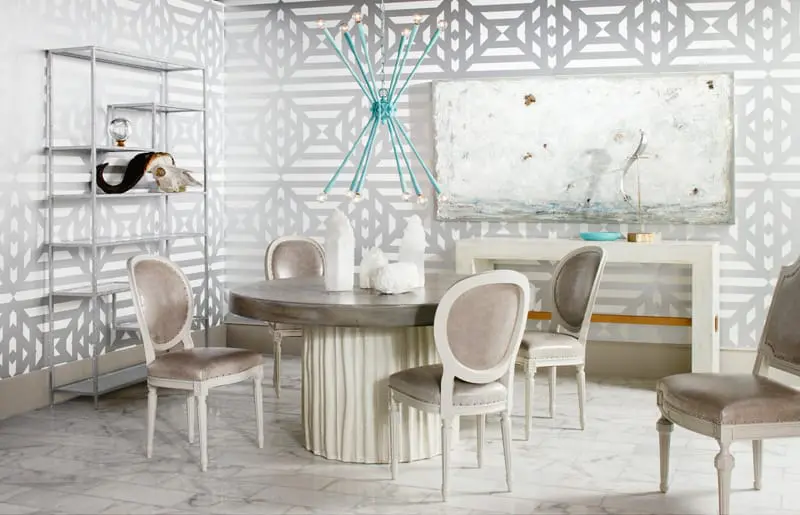 Dining room with a round concrete table, upholstered chairs, a silver shelving unit, and abstract art on a geometric patterned wall.