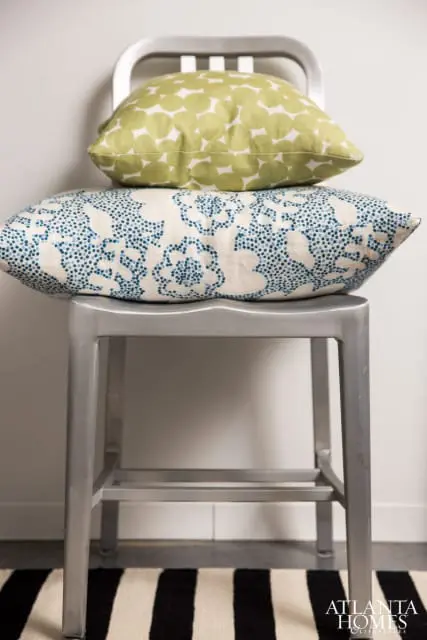 Two decorative pillows stacked on a metal stool. The top pillow is lime green with a white circular pattern. The bottom pillow is cream with a blue dotted floral pattern.
