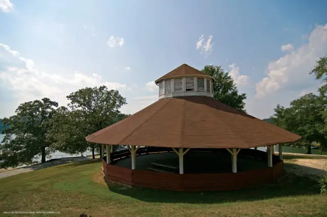 Octagonal wooden pavilion with a brown shingled roof and white support posts, set on a grassy hill overlooking a lake and surrounded by trees.