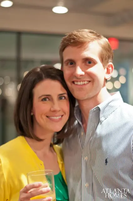 A woman in a yellow cardigan and a man in a striped button-down shirt smile at the camera. The woman holds a glass of a light-colored beverage.