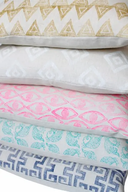 Stack of five decorative pillows with various patterns: gold triangles, white diamonds, pink eyes, turquoise fish, and blue Greek key.