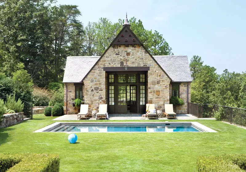 Stone cottage with a steep gabled roof, large glass doors, and a swimming pool in the manicured lawn.