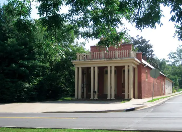 Red brick building with a covered porch supported by tall, cream-colored columns and a white railing on the roofline, surrounded by lush green trees.