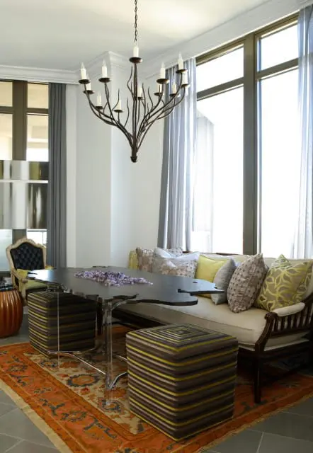 Dining area with a dark wood live-edge table, acrylic legs, upholstered banquette seating with patterned pillows, striped ottomans, and a wrought iron chandelier.
