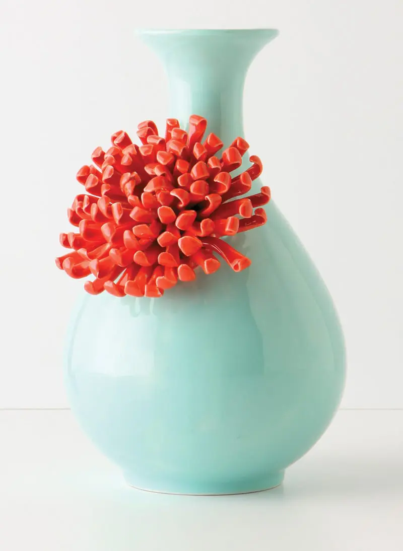 Close-up of a light blue ceramic vase with a bright orange, spiky floral accent attached to its side.