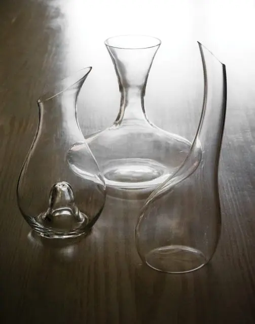 Three clear glass wine decanters with curved spouts and wide bases sit on a dark wood surface.