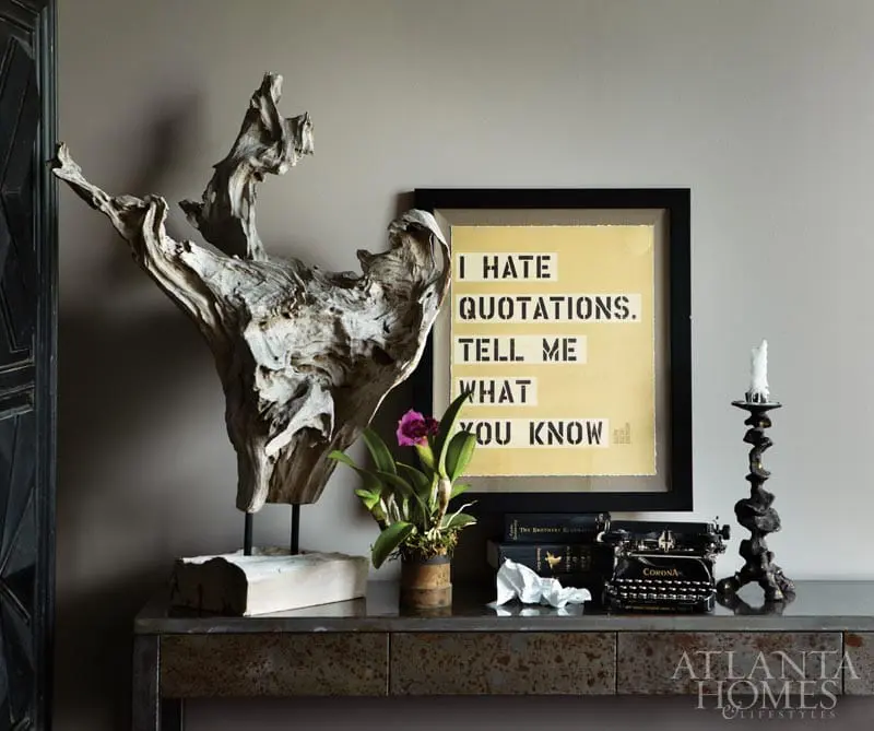 A vignette on a metal console table features a large driftwood sculpture, a framed yellow print with black text reading "I HATE QUOTATIONS. TELL ME WHAT YOU KNOW", a potted orchid, a vintage typewriter, and a black candlestick with a white candle.