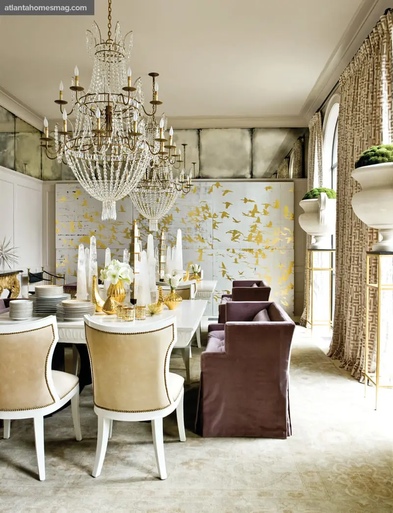 Elegant dining room with two crystal chandeliers, a white table set with gold accents, upholstered chairs in cream and plum, and a wall mural of gold birds in flight.