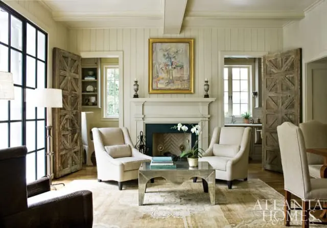 Living room with a fireplace, two cream upholstered armchairs, a mirrored coffee table, and rustic wooden doors.