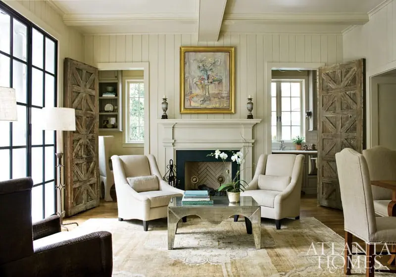 Living room with a fireplace, two cream upholstered armchairs, a mirrored coffee table, and rustic wooden doors.