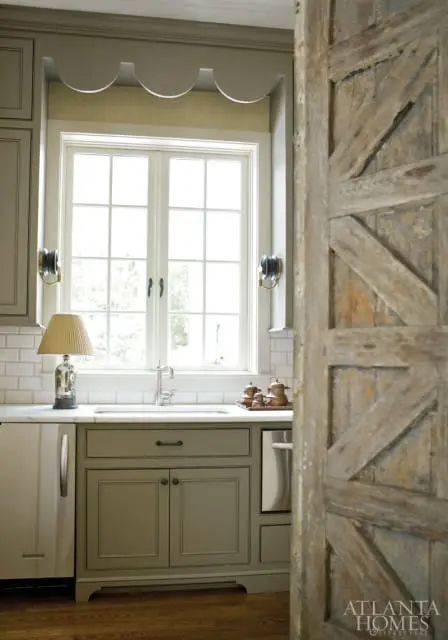 Kitchen with sage green cabinetry, white subway tile backsplash, and a double-hung window above the sink. A rustic wooden barn door is visible on the right.