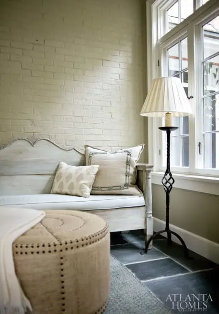 Light green painted brick wall behind a pale blue distressed wood bench with decorative pillows and a round upholstered ottoman. A floor lamp stands next to a window.