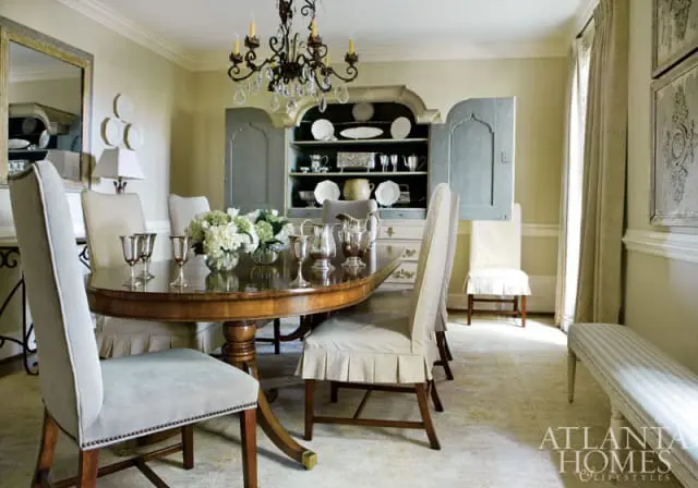 Dining room with oval wooden table set with floral arrangement and silverware, upholstered chairs, ornate chandelier, and a large gray hutch filled with dishes.