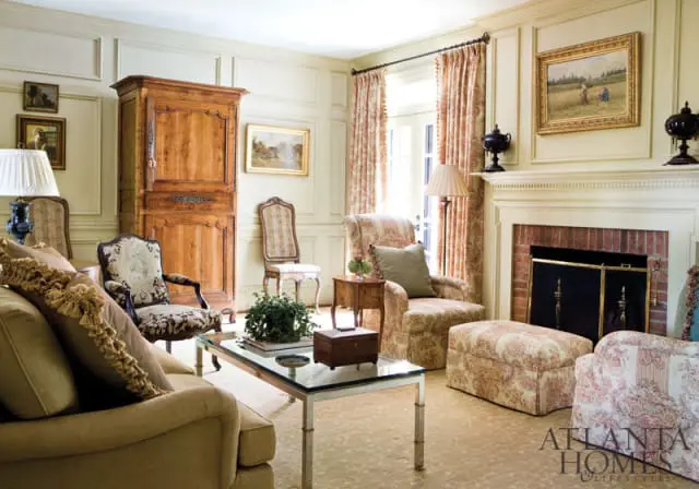 Living room with paneled walls, a large wooden armoire, floral upholstered furniture, a glass-top coffee table, and a brick fireplace with a white mantel.