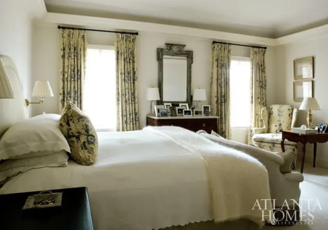 Bedroom with white bedding, patterned curtains, a dark wood dresser with framed photos, and a wingback chair.
