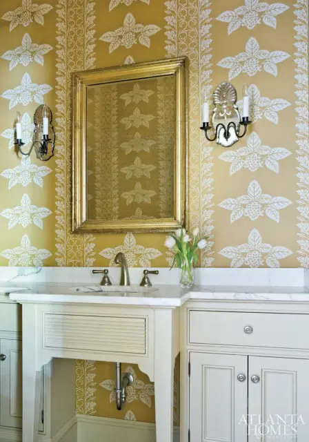 Powder room with gold floral wallpaper, a gold framed mirror, and a white vanity with a marble countertop and brass fixtures.
