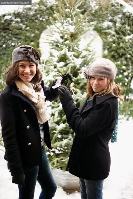 Two smiling women in winter coats and hats stand in front of a snow-covered evergreen tree, holding mistletoe.