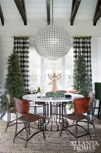 Dining room with white shiplap walls, dark wood beams, and a large spherical pendant light. A round marble-top table is set with leather chairs, Christmas trees, and holiday decor.