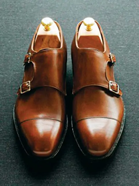 Pair of brown leather double monk strap shoes with cap toes and brass buckles, displayed on shoe trees.
