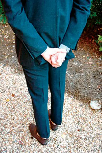 Man in a dark suit with hands clasped behind his back, standing on a gravel path with some dry leaves and greenery in the background.