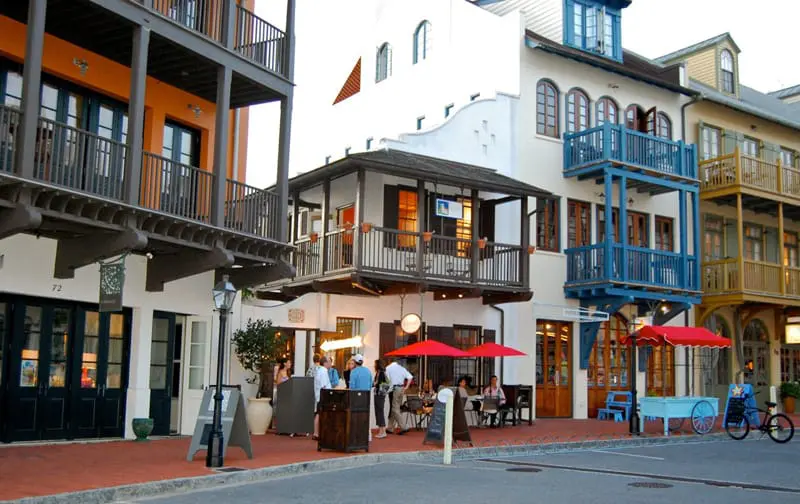 Row of buildings with balconies, some painted orange, white, and blue, with outdoor seating and red umbrellas in front of shops.