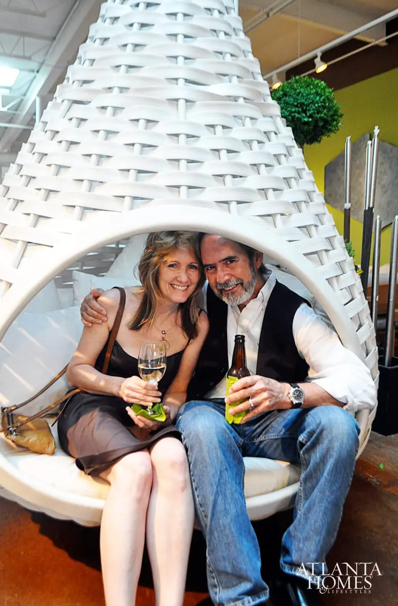 Couple sitting in a white woven pod chair, woman holding a wine glass, man holding a beer bottle. Atlanta Homes & Lifestyles logo in the bottom right corner.