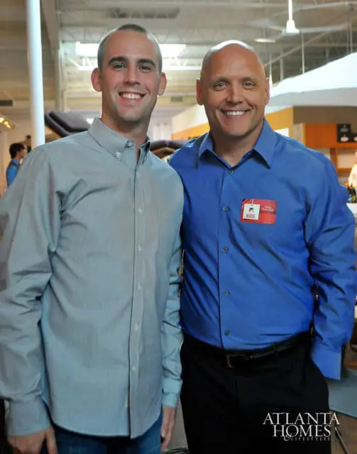 Two men smiling at the camera. The man on the left wears a light blue button-down shirt and jeans. The man on the right wears a royal blue button-down shirt with a red "MADE" name tag and black pants.