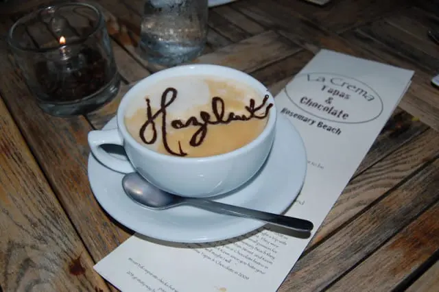 White coffee cup with latte art spelling "Hazelnut" in chocolate drizzle, served on a saucer with a spoon, next to a menu for La Crema Tapas & Chocolate in Rosemary Beach.
