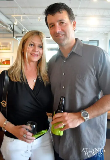 A blonde woman in a black top and white pants holds a wine glass, while a man in a gray shirt holds a bottle of beer and a green napkin.