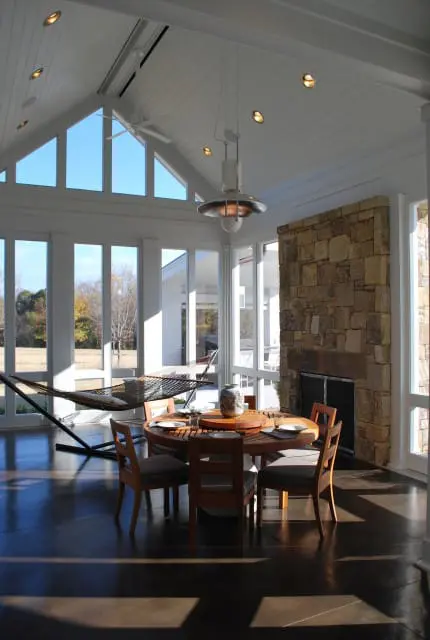 Sunroom with a round wooden dining table, chairs, stone fireplace, and a hammock by large windows overlooking a yard.