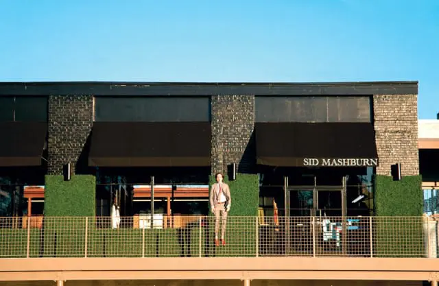 Building with dark brick facade, black awnings, and a mannequin dressed in a tan suit standing on a balcony with green hedges and a metal fence.