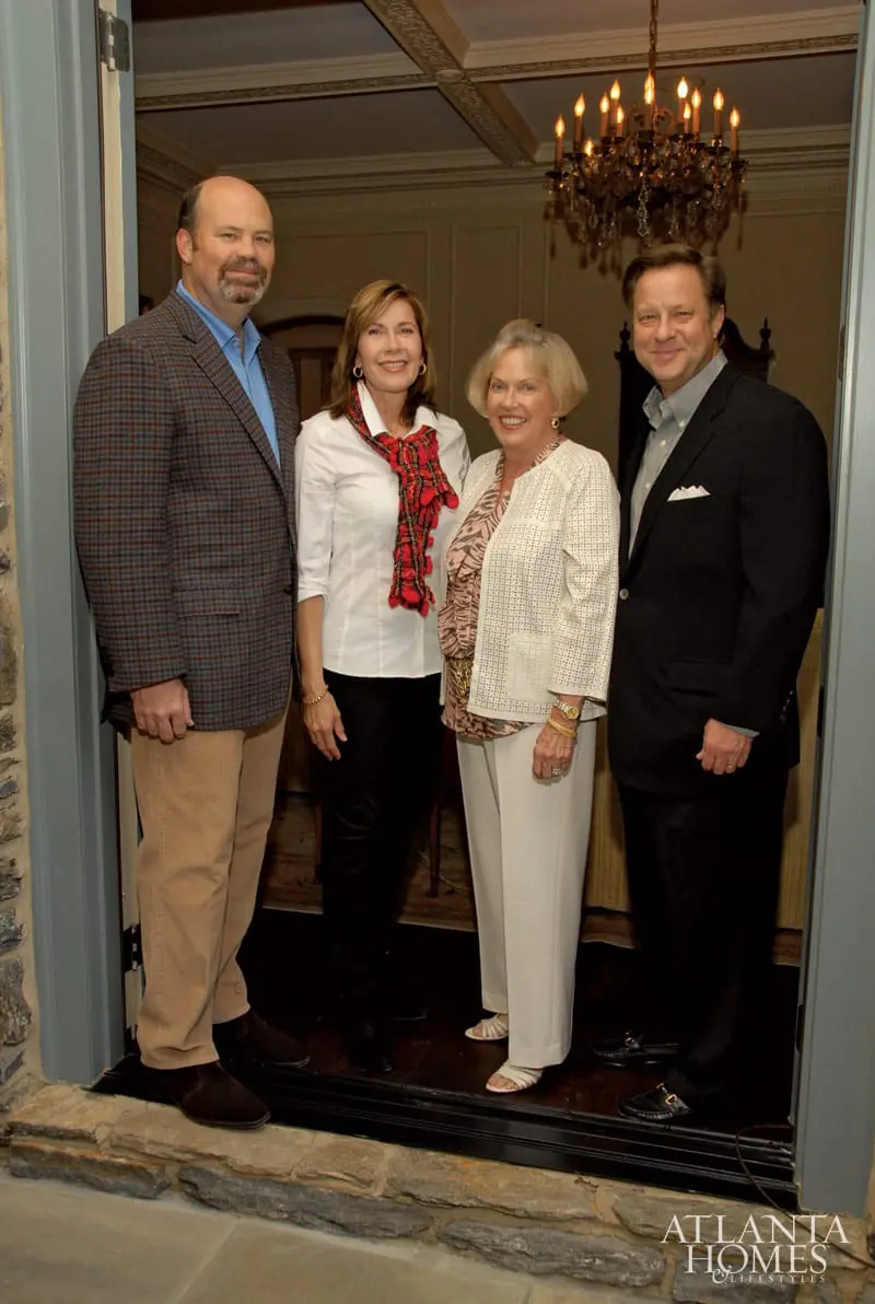 Four people stand in a doorway. A man in a plaid jacket and blue shirt is on the left. Next to him is a woman in a white shirt and red scarf. A woman in a cream jacket and patterned blouse stands in the center. A man in a black suit and gray shirt is on the right.