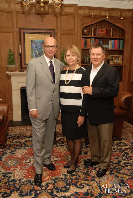 Three people stand in a room with wood-paneled walls and a large oriental rug. A man in a grey suit stands on the left, a woman in a black and white striped top and black skirt stands in the middle, and a man in a black blazer and khaki pants stands on the right.
