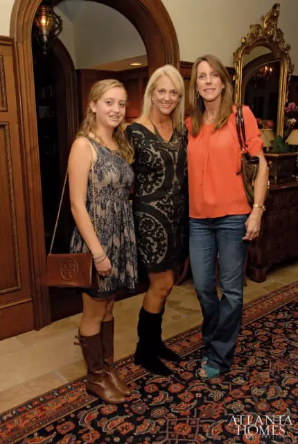 Three women standing in a room with wood paneling, a large ornate mirror, and a patterned rug. The woman on the left wears a blue lace dress and brown boots. The middle woman wears a black lace dress and statement necklace. The woman on the right wears an orange top and blue jeans.