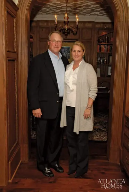 Man in a dark suit and blue collared shirt and woman in a white ruffled blouse and gray cardigan stand in a wood-paneled room with bookshelves and a chandelier.