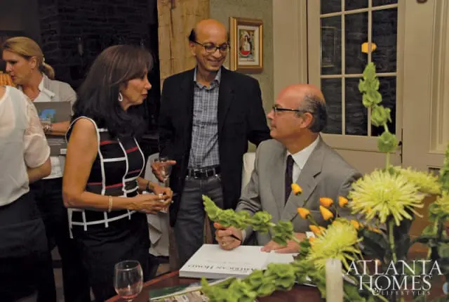 Three people at a table with a book and floral arrangements. A man in a suit signs a book titled "AMERICAN HOMES". The Atlanta Homes & Lifestyles logo is visible in the lower right.