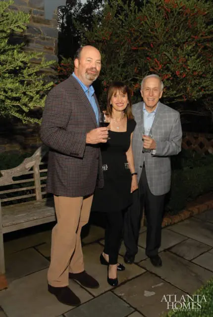 Three people stand outdoors on a stone patio. A man in a plaid jacket and tan pants holds a wine glass. A woman in a black dress stands between him and another man in a gray plaid jacket.