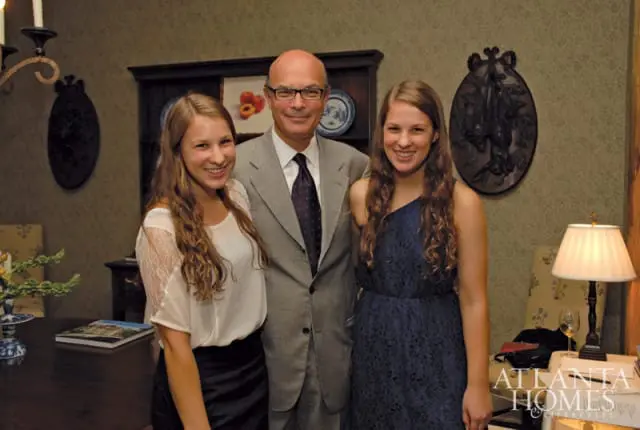 Man in a gray suit and purple tie stands between two young women, one in a white lace top and black skirt, the other in a navy blue dress, in a room with patterned wallpaper and dark wood furniture.