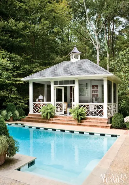Screened-in porch with white lattice and a cupola sits beside a bright blue swimming pool surrounded by lush green trees.