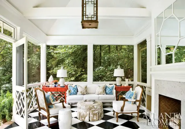 Screened porch with black and white checkered floor, white sofa, two orange console tables, upholstered chairs, and a floral ottoman. Lush green trees are visible through the windows.