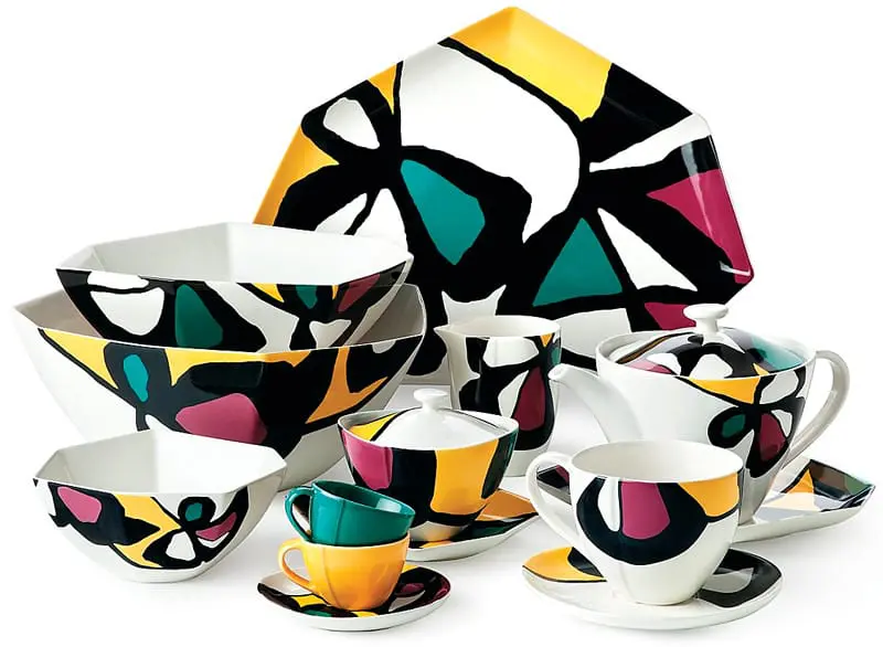 Collection of white ceramic tableware featuring a bold, abstract geometric pattern in black, yellow, teal, and magenta. Includes bowls, cups, saucers, a teapot, and a large platter.