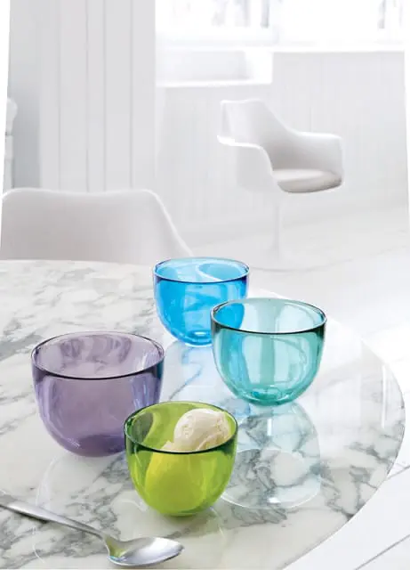 Four colorful glass bowls in purple, green, turquoise, and blue sit on a marble tabletop. The green bowl contains ice cream.