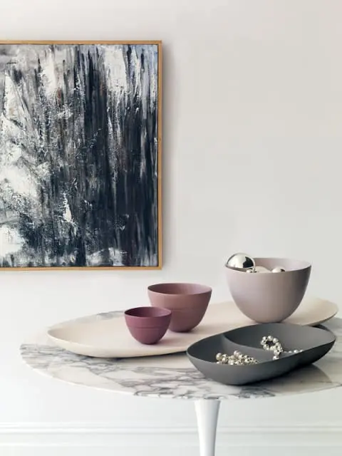 Abstract art with black, white, and gray brushstrokes hangs above a marble-topped table displaying various decorative bowls and trays in muted tones of pink, gray, and cream.