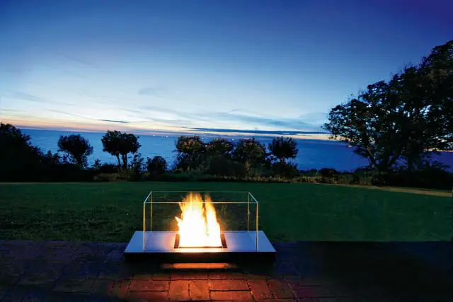Modern outdoor fireplace with glass enclosure on brick patio overlooking the ocean at dusk with lush green lawn and trees