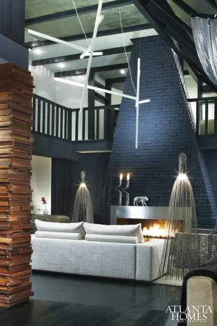 Modern living room with a dark gray brick fireplace, light gray sofa, and a sculptural wooden wall feature. A geometric chandelier hangs from the ceiling.