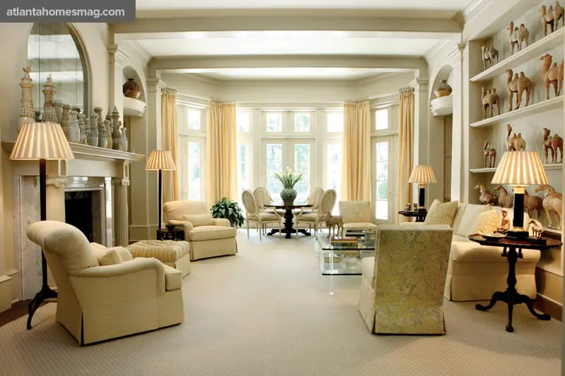 Cream-colored living room with a fireplace, bay windows, plush armchairs, a glass coffee table, and shelves displaying decorative figurines.