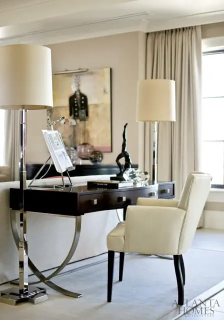 Cream upholstered armchair next to a dark wood desk with chrome accents, featuring two lamps, artwork, and decorative objects.