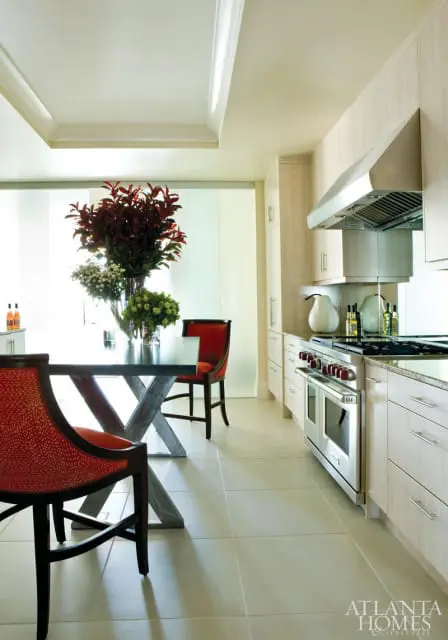 Modern kitchen with light wood cabinetry, stainless steel range, and a dining area with red upholstered chairs and a dark metal table with a large floral arrangement.