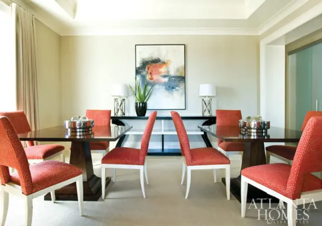 Dining room with two dark wood tables, each with four chairs upholstered in red patterned fabric. A console table with abstract art and two lamps is behind the tables.